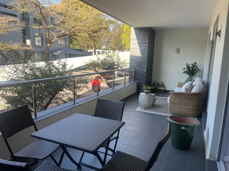 A spacious covered balcony featuring modern tiled flooring, a stainless steel balustrade, and a decorative stone feature wall, offering a pleasant outdoor seating area.