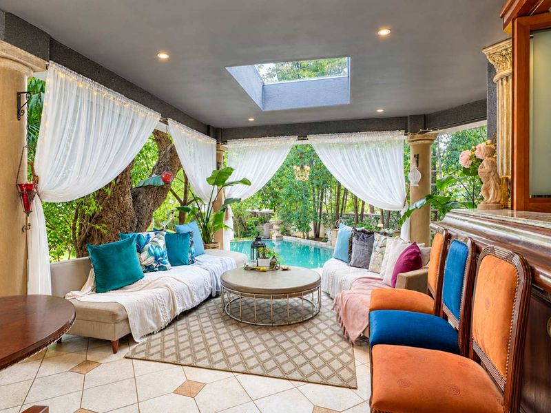 A covered outdoor patio area featuring a skylight, stone columns, tiled flooring, and a built-in bar, overlooking a swimming pool and garden.