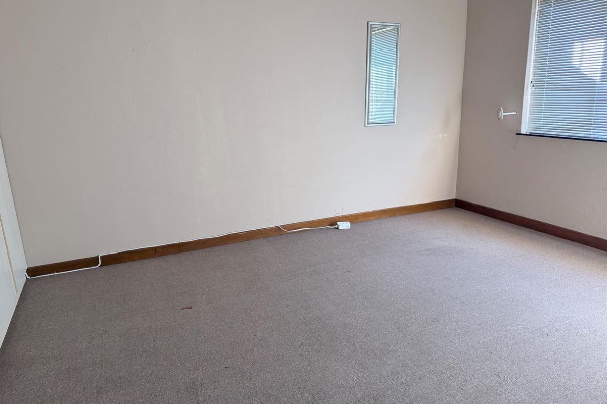 An empty bedroom featuring neutral wall tones, carpeted flooring, timber skirting boards, and a window fitted with venetian blinds.