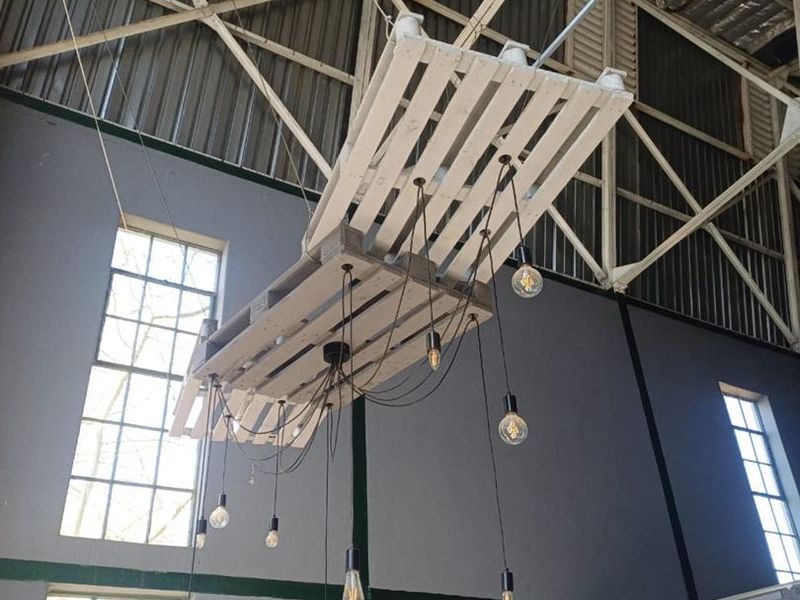 Spacious industrial interior featuring high ceilings with exposed steel trusses, large windows providing natural light, and a unique custom pallet pendant light fixture.