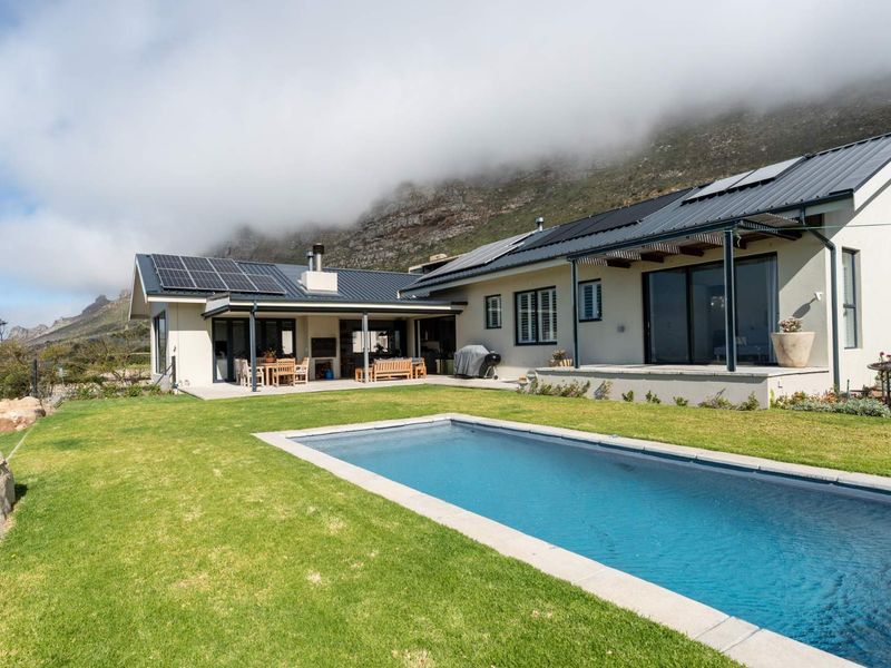 Rear view of a modern home featuring a swimming pool, covered patio with built-in braai, and solar panels set against a mountain backdrop.