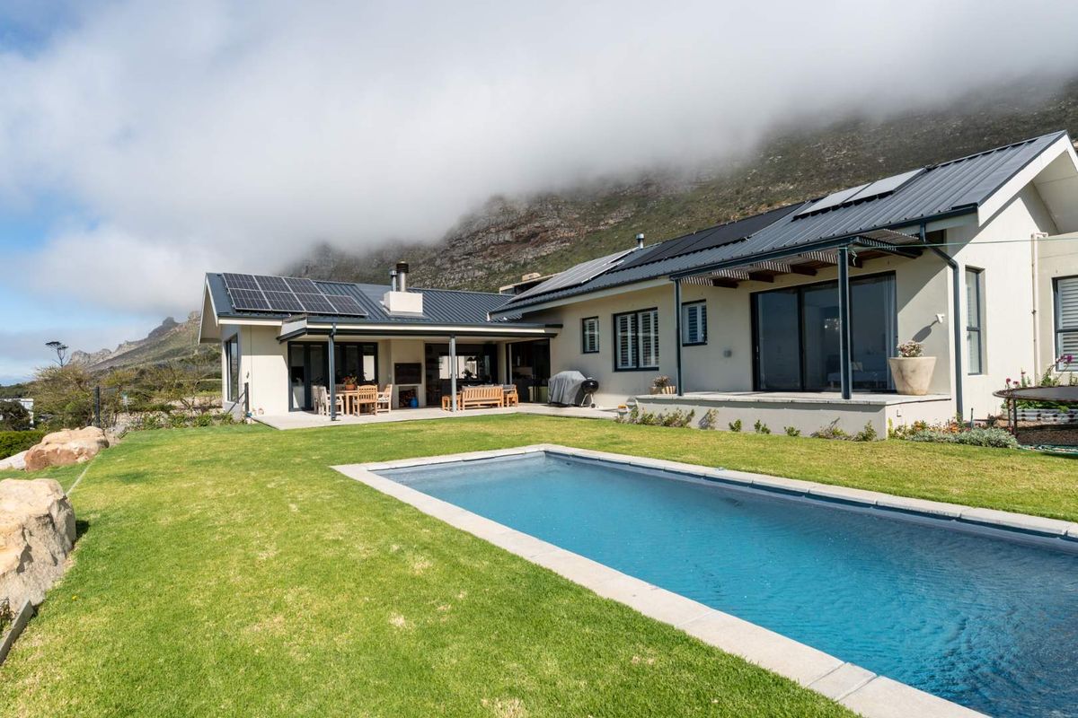 Rear view of a modern home featuring a swimming pool, covered patio with built-in braai, and solar panels set against a mountain backdrop.