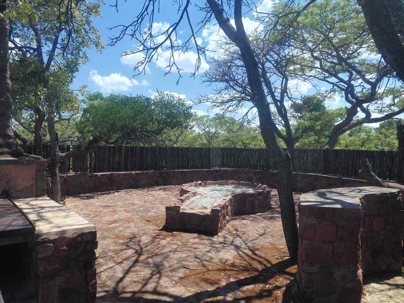 Outdoor entertainment area featuring a stone-built fire pit and integrated braai, set within a natural garden environment with timber fencing.