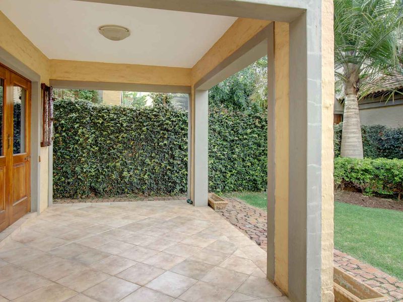 A covered patio area featuring tiled flooring, concrete structural pillars, and wooden double doors leading into the home, overlooking a private garden.