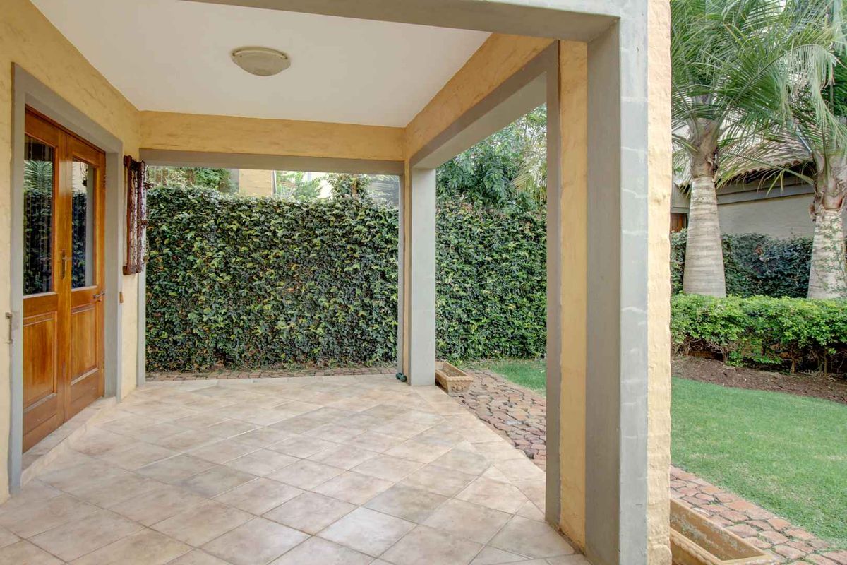 A covered patio area featuring tiled flooring, concrete structural pillars, and wooden double doors leading into the home, overlooking a private garden.