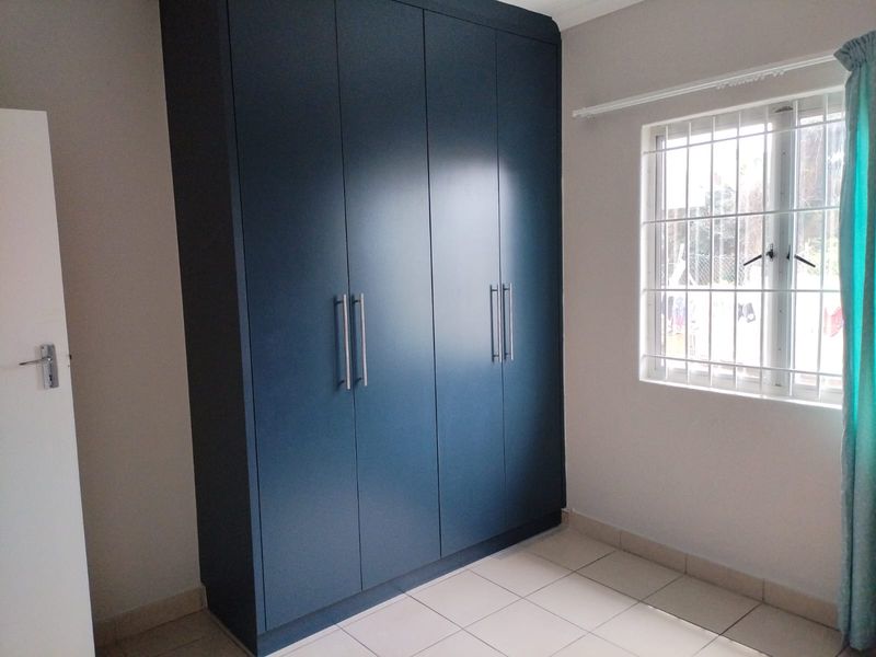 A clean, empty bedroom featuring modern dark blue built-in cupboards, neutral tiled flooring, and a window fitted with security bars.