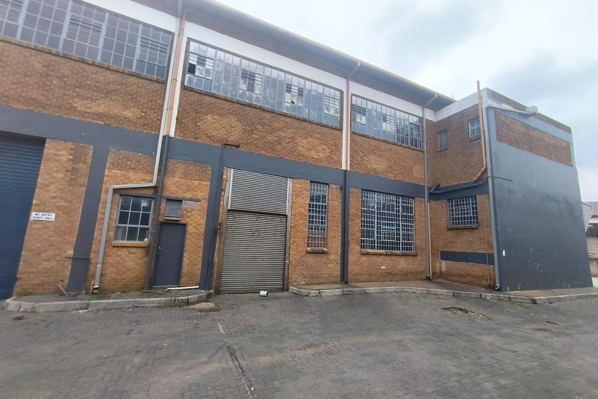 Industrial warehouse exterior featuring brick facade, multiple steel-framed windows, and roller shutter access doors, showing signs of wear and maintenance requirements.