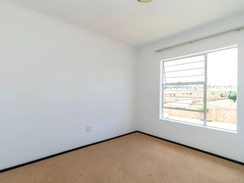 A bright, empty bedroom featuring neutral carpet flooring, white walls, and a large window with security bars.