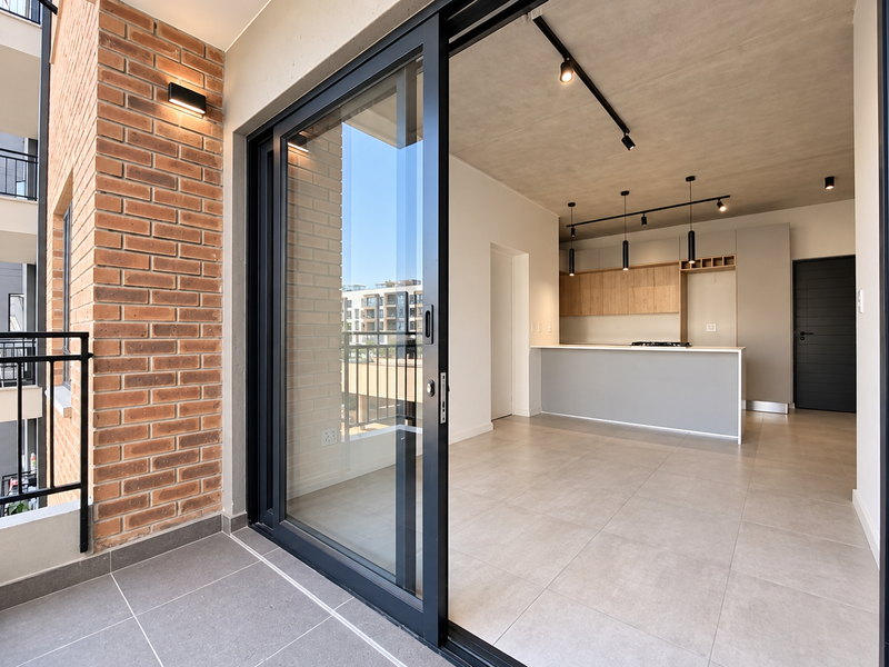 Modern open-plan apartment featuring a seamless transition to a private balcony, exposed concrete ceilings, track lighting, and a contemporary kitchen with built-in cabinetry.