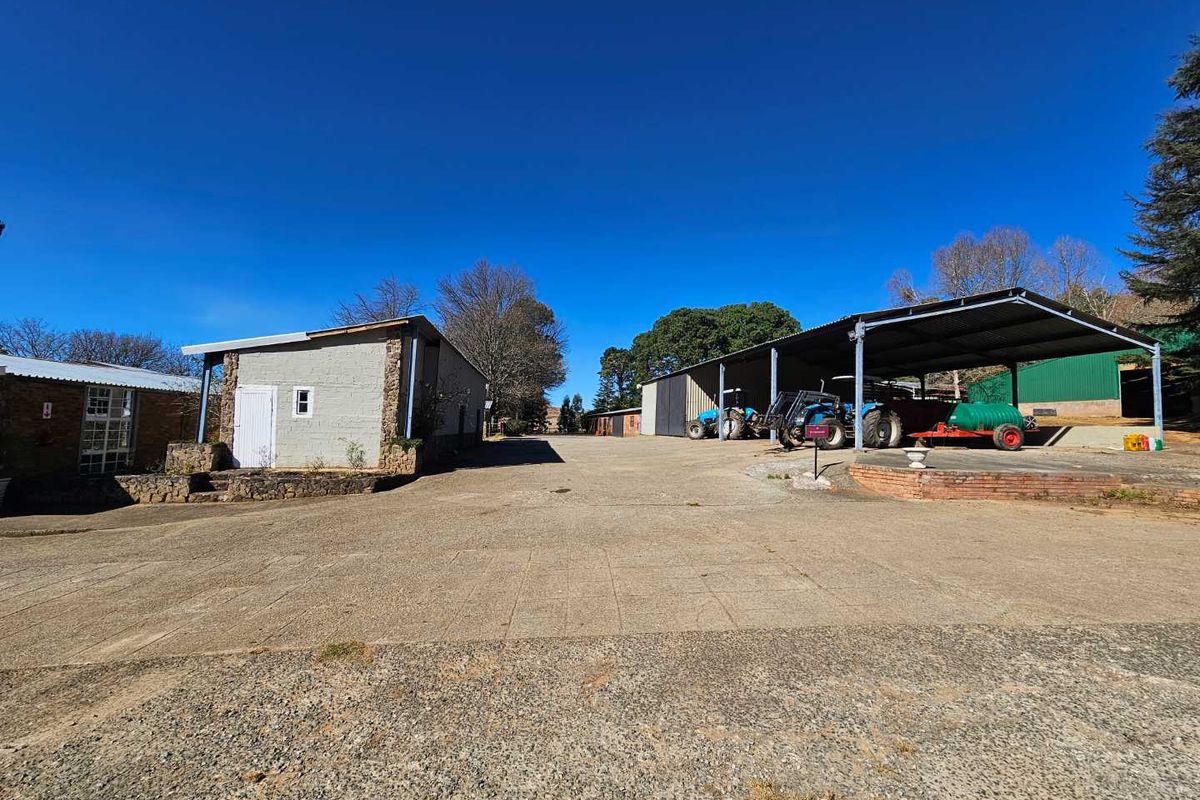 A spacious farm yard featuring a large concrete driveway, a detached outbuilding, and a covered machinery storage shed for agricultural equipment.