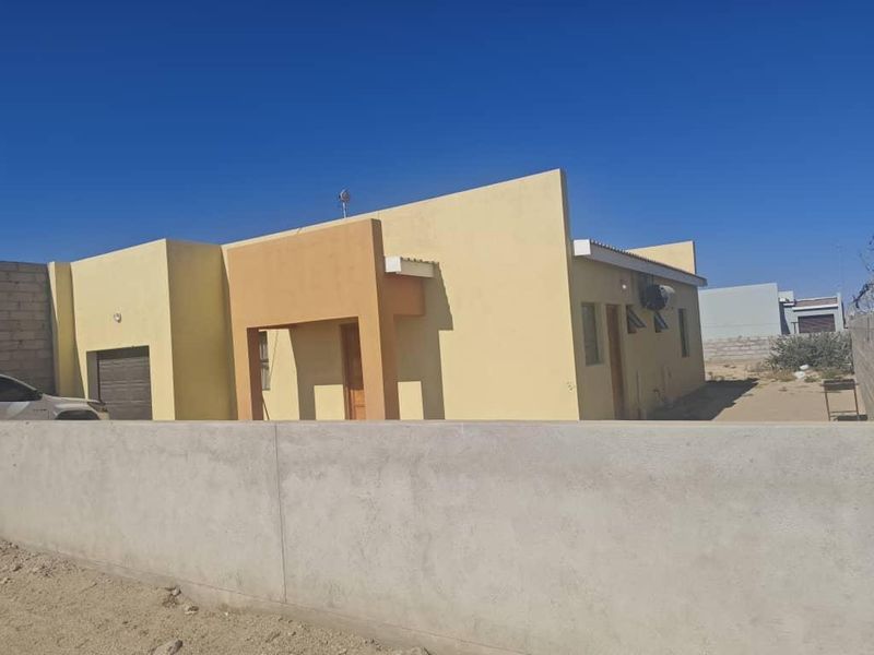 Front exterior view of a single-story house featuring a painted facade, integrated garage, and a concrete boundary wall on an unpaved lot.