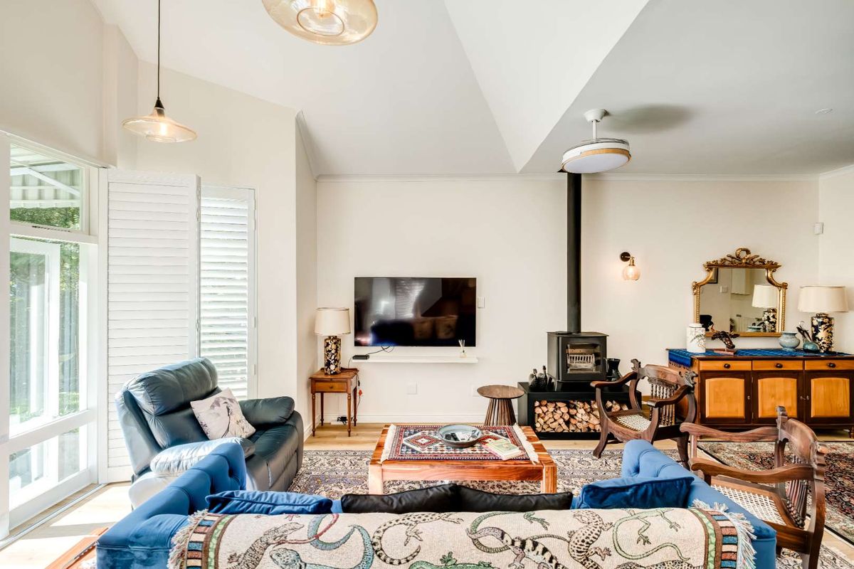 A bright living room featuring a vaulted ceiling, a built-in wood-burning fireplace, and large windows with plantation shutters providing natural light.