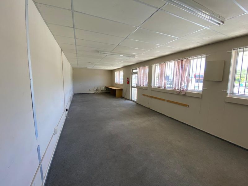 An empty, open-plan office space featuring a suspended ceiling, fluorescent lighting, carpeted flooring, and security bars on the windows.
