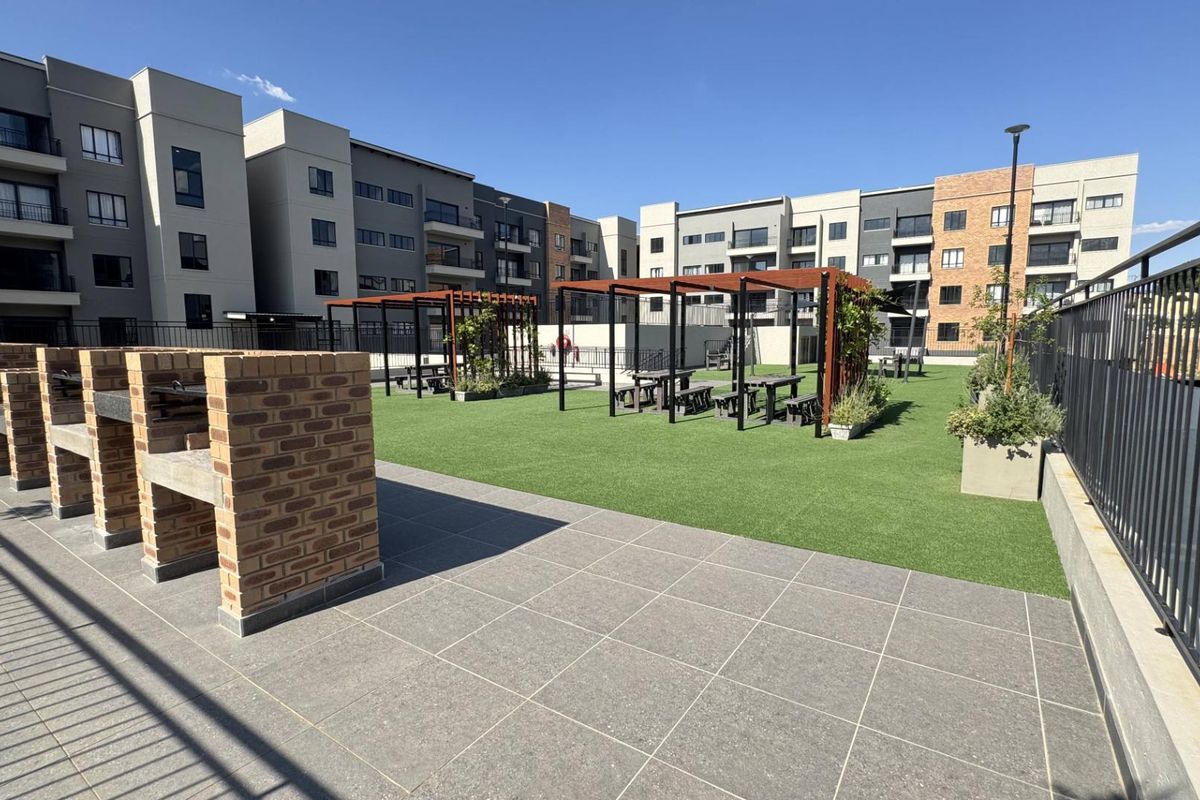 Communal outdoor entertainment area featuring brick-built braai stations, artificial lawn, and shaded pergola seating areas within a modern apartment complex.