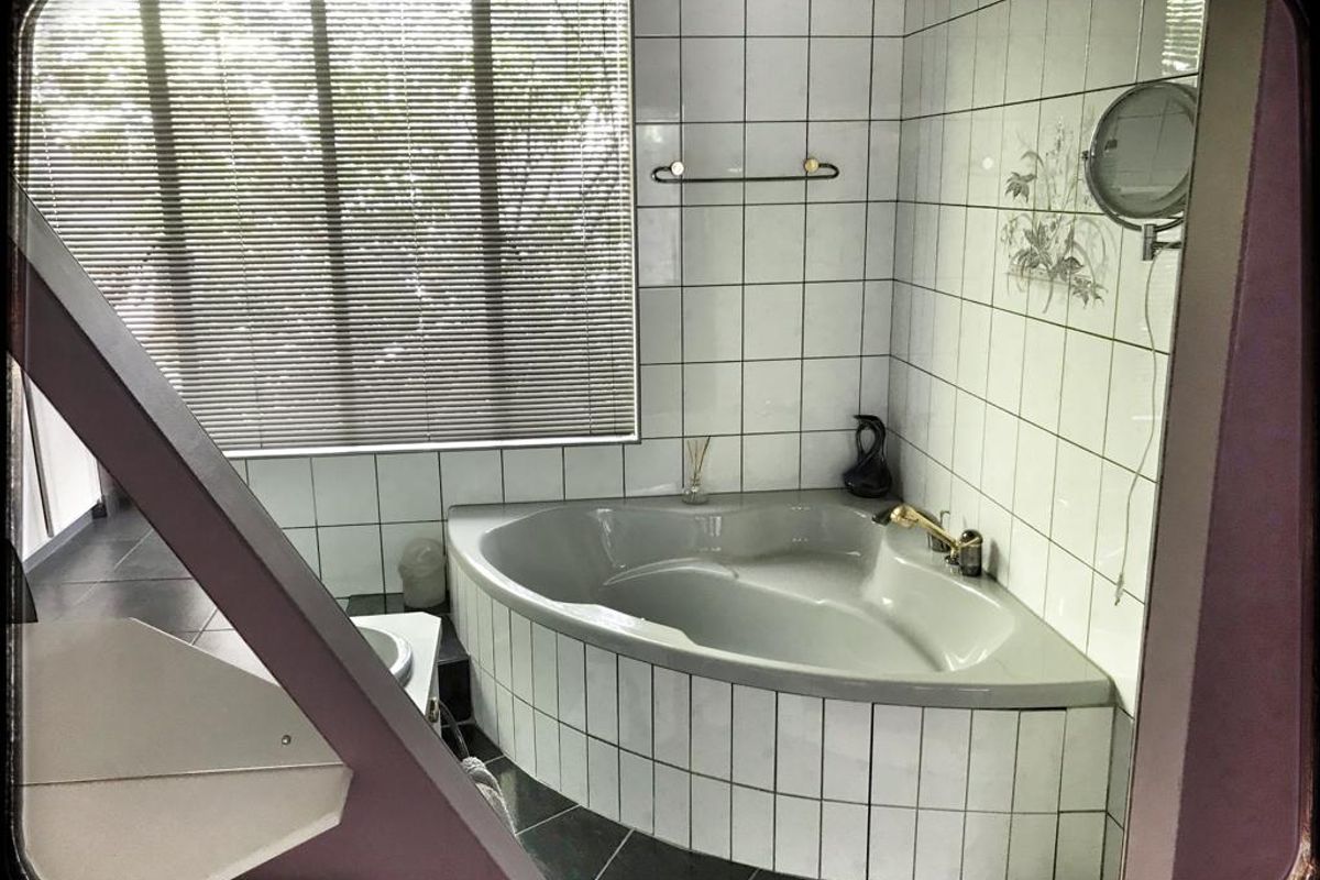 A bathroom featuring a corner bathtub with white wall tiles, dark floor tiles, and a window with blinds.