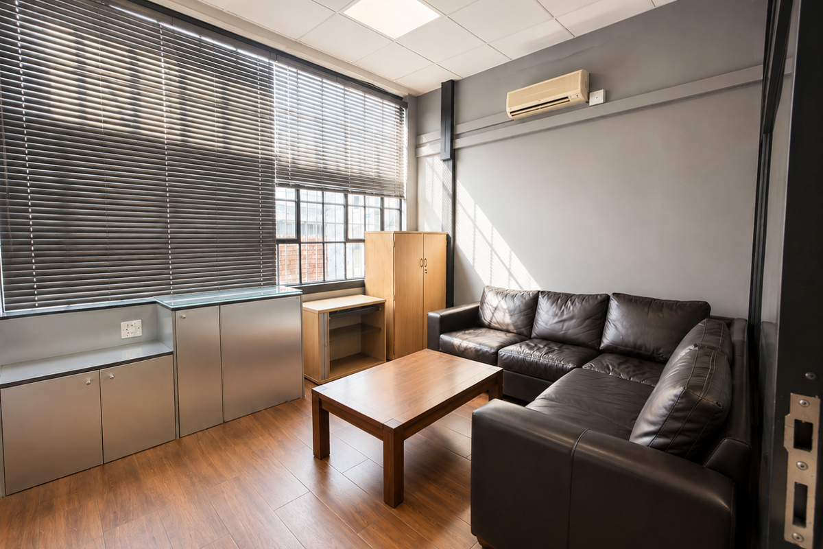 A functional office or lounge space featuring laminate flooring, built-in storage cabinetry, large windows with venetian blinds, and a wall-mounted air conditioning unit.