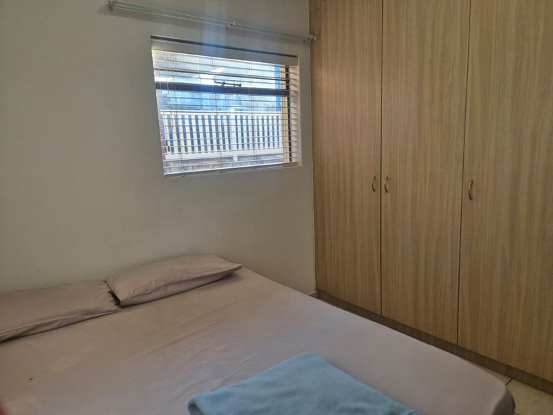A functional bedroom featuring light wood-finish built-in cupboards and a window with fitted venetian blinds.