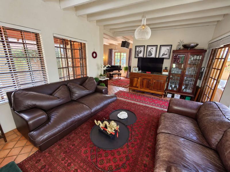 A spacious living room featuring exposed ceiling beams, terracotta floor tiles, and large windows with venetian blinds, offering a warm and traditional atmosphere.