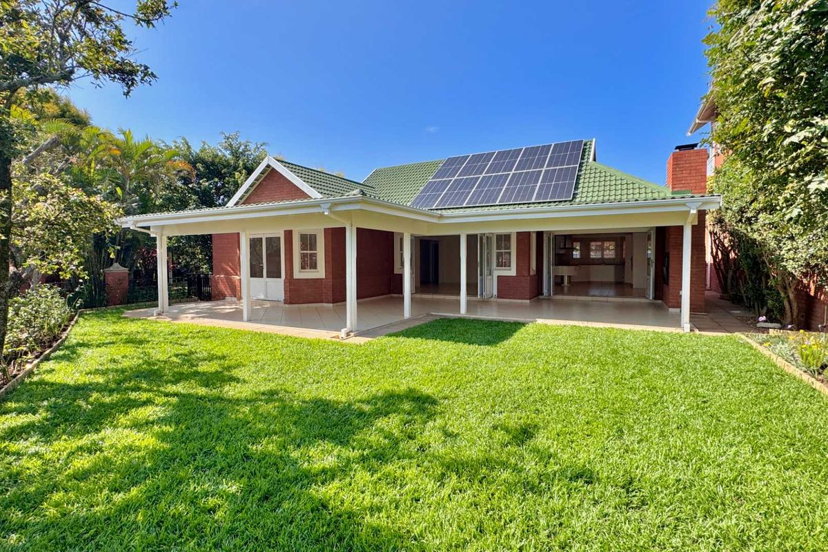 Rear view of a brick house featuring a covered patio, green tiled roof with solar panels, and a well-maintained garden.