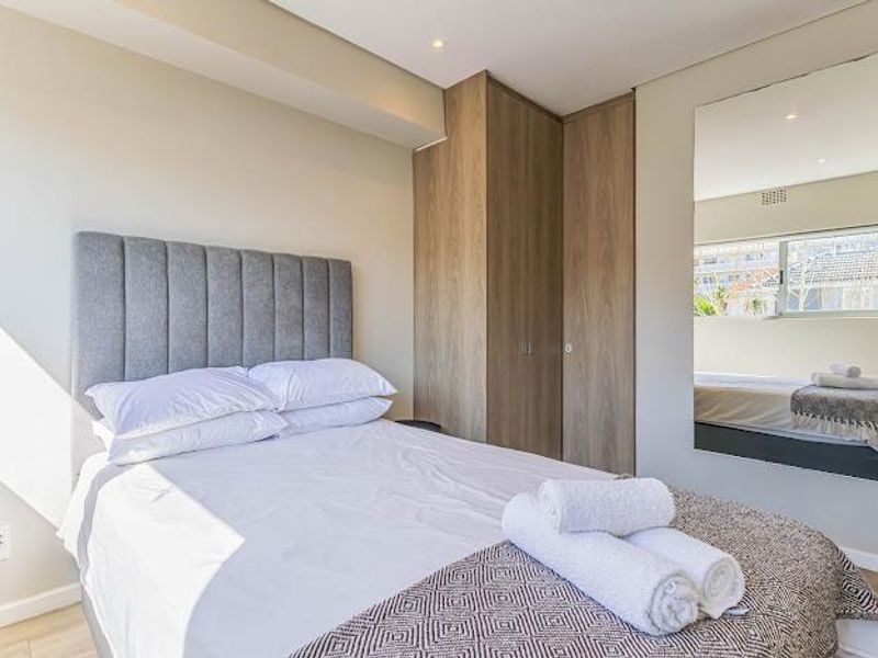 A well-maintained bedroom featuring built-in wooden cupboards, recessed ceiling lighting, and a large wall-mounted mirror reflecting the window.