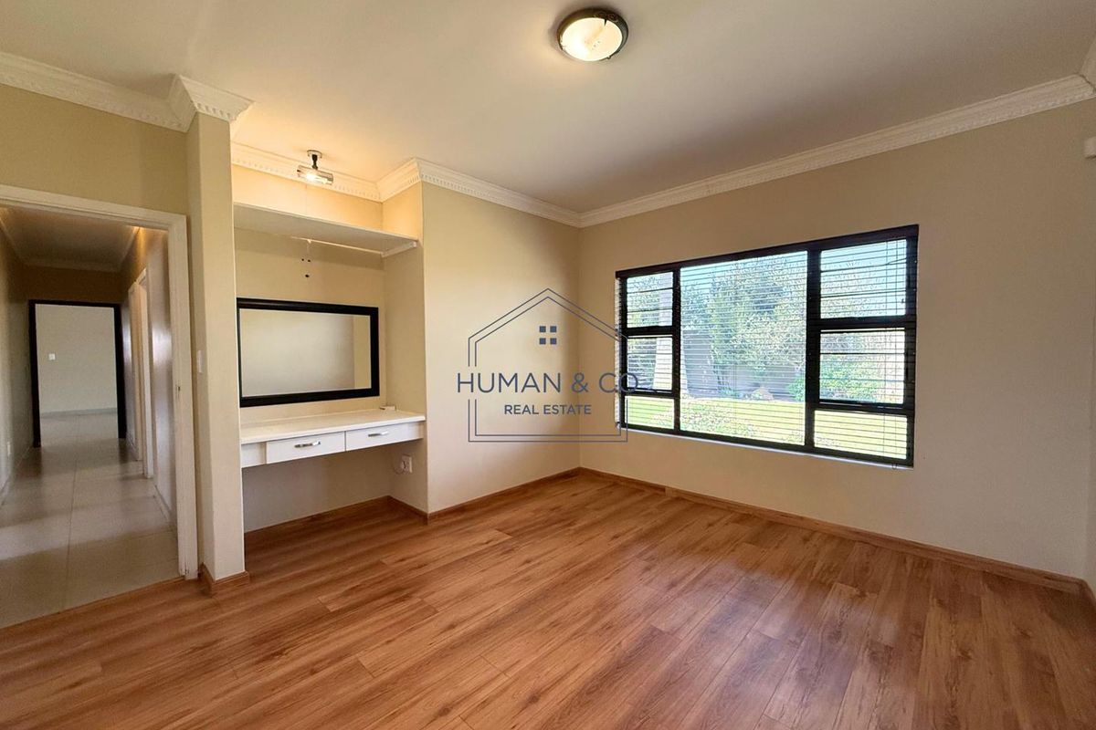 A bright bedroom featuring wood-look flooring, a built-in vanity station with mirror, and large windows overlooking the garden.