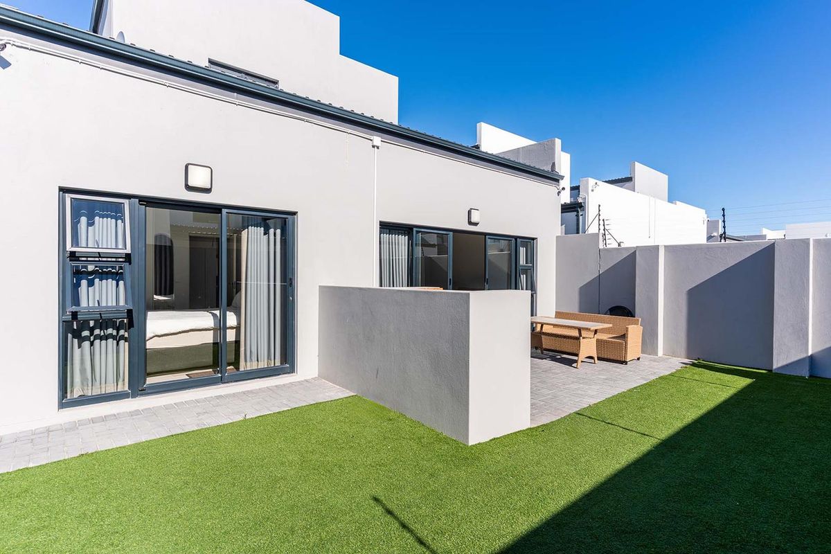 A modern rear exterior view featuring a paved patio area, artificial lawn, and sliding glass doors providing access to the interior living space.