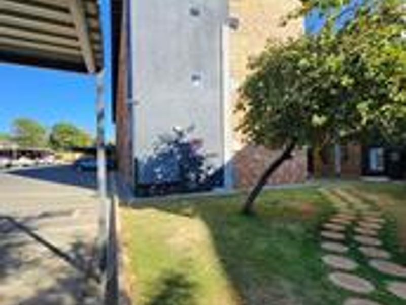 Exterior view of the apartment building featuring a brick and plastered facade, adjacent carport, and a landscaped garden area with a paved walkway.