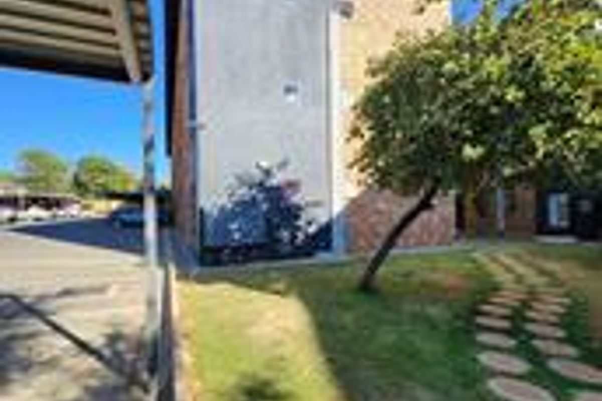 Exterior view of the apartment building featuring a brick and plastered facade, adjacent carport, and a landscaped garden area with a paved walkway.
