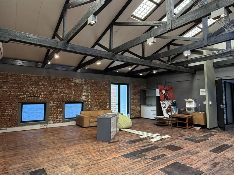Spacious industrial-style office featuring exposed timber roof trusses, skylights, an exposed brick feature wall, and hardwood flooring with integrated kitchenette cabinetry.