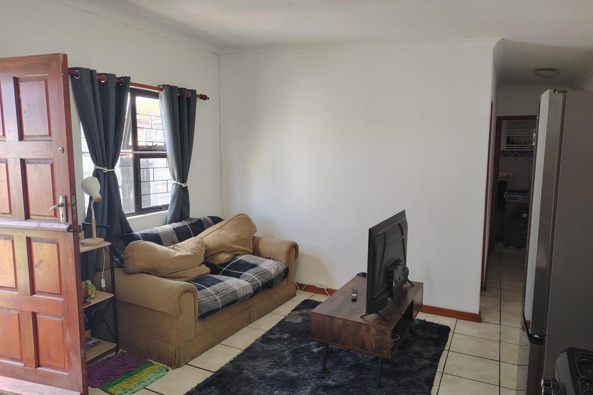 A compact living area featuring tiled flooring, a timber entrance door, and a window with security bars, leading through to a laundry area.