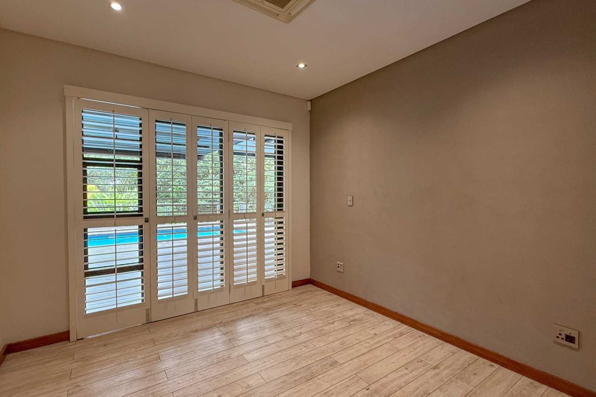 A bright, empty room featuring light wood-look flooring, recessed ceiling lighting, and white plantation shutters opening to an outdoor pool area.