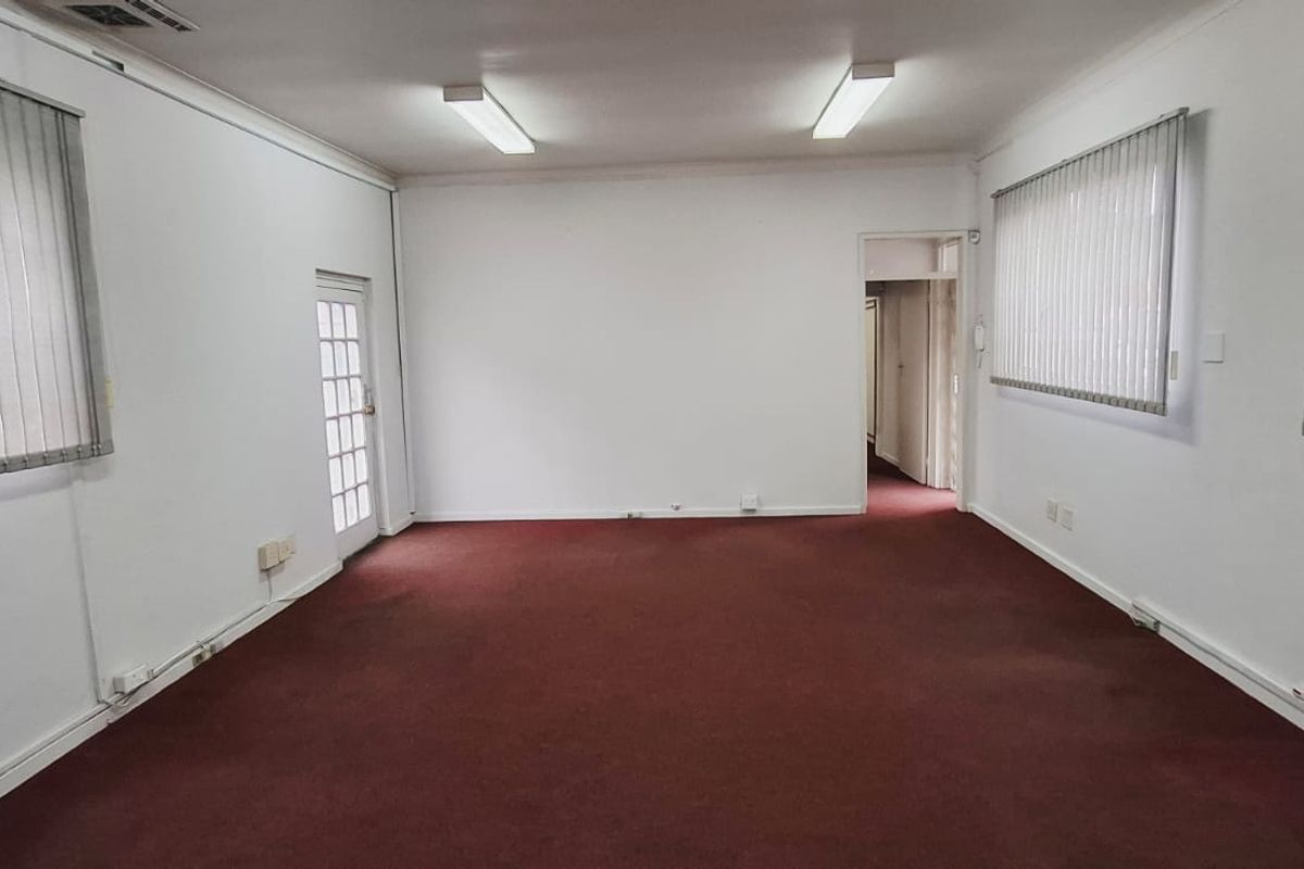 A spacious, empty office area featuring neutral walls, carpeted flooring, vertical blinds, and fluorescent lighting, suitable for a professional workspace.