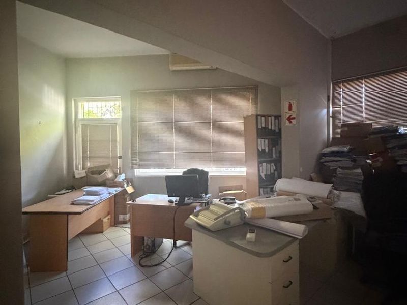 A functional office space featuring tiled flooring, air conditioning, and large windows with venetian blinds, currently utilized for storage and workspace.