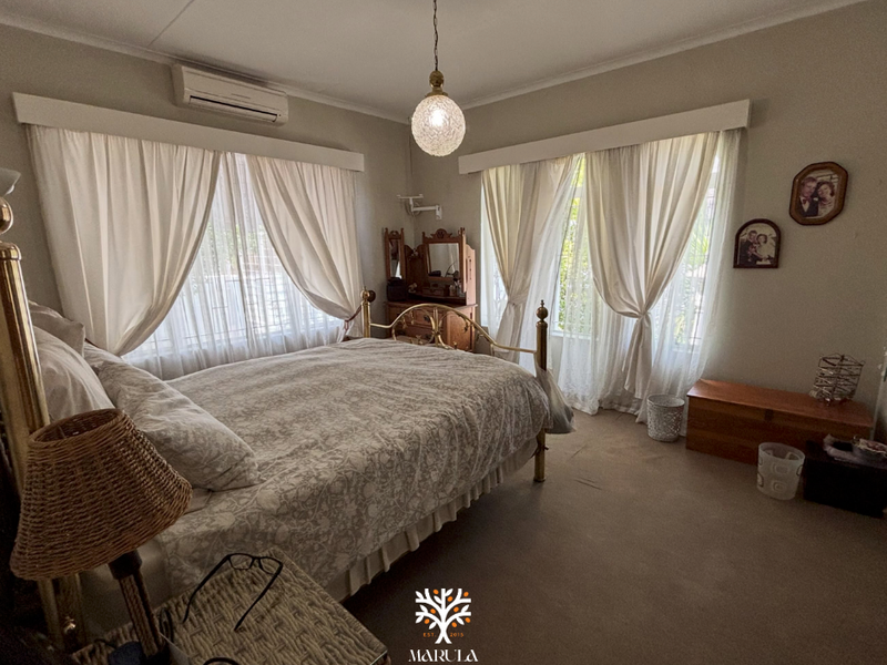 A spacious bedroom featuring neutral walls, carpeted flooring, a wall-mounted air conditioning unit, and dual windows with curtain treatments.