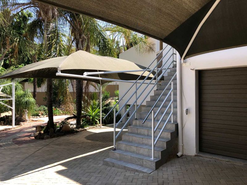 Rear exterior area featuring a paved driveway, a tiled staircase with metal handrails, and overhead shade sails providing cover.