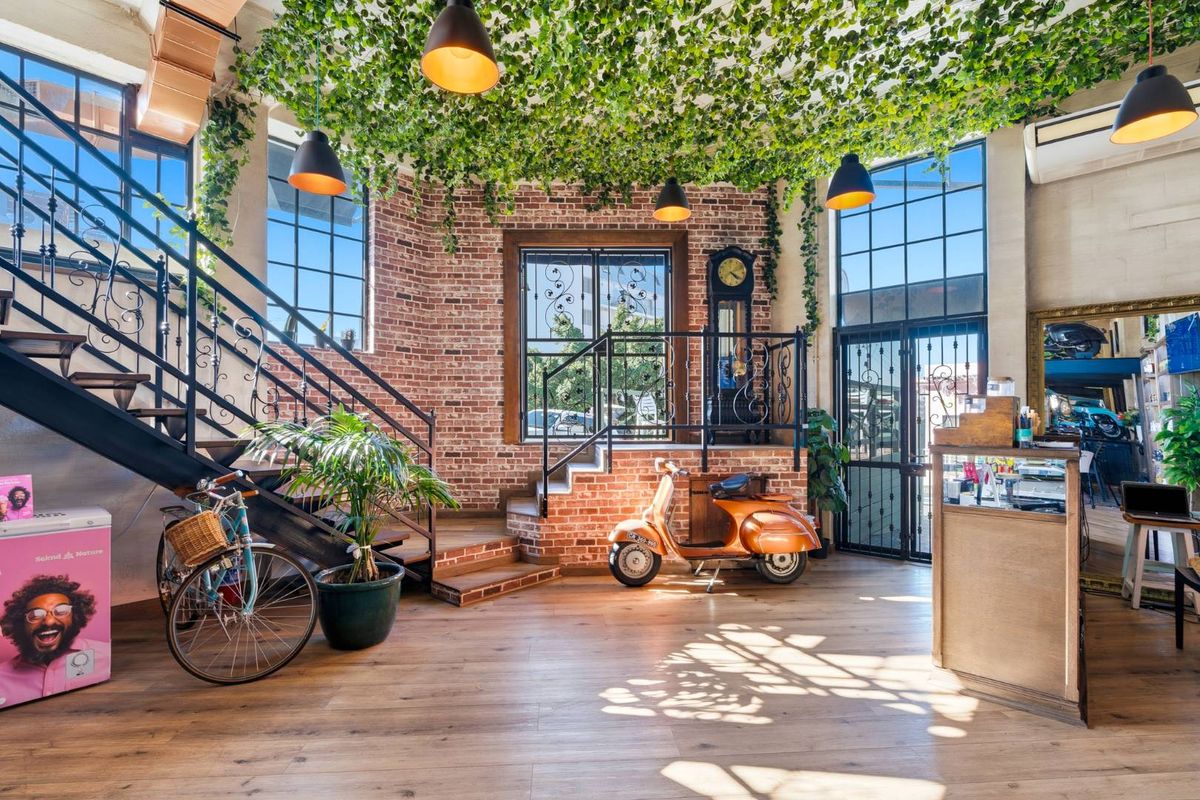 A stylish retail interior featuring exposed brick walls, large industrial-style windows, a feature staircase, and high ceilings with decorative greenery.