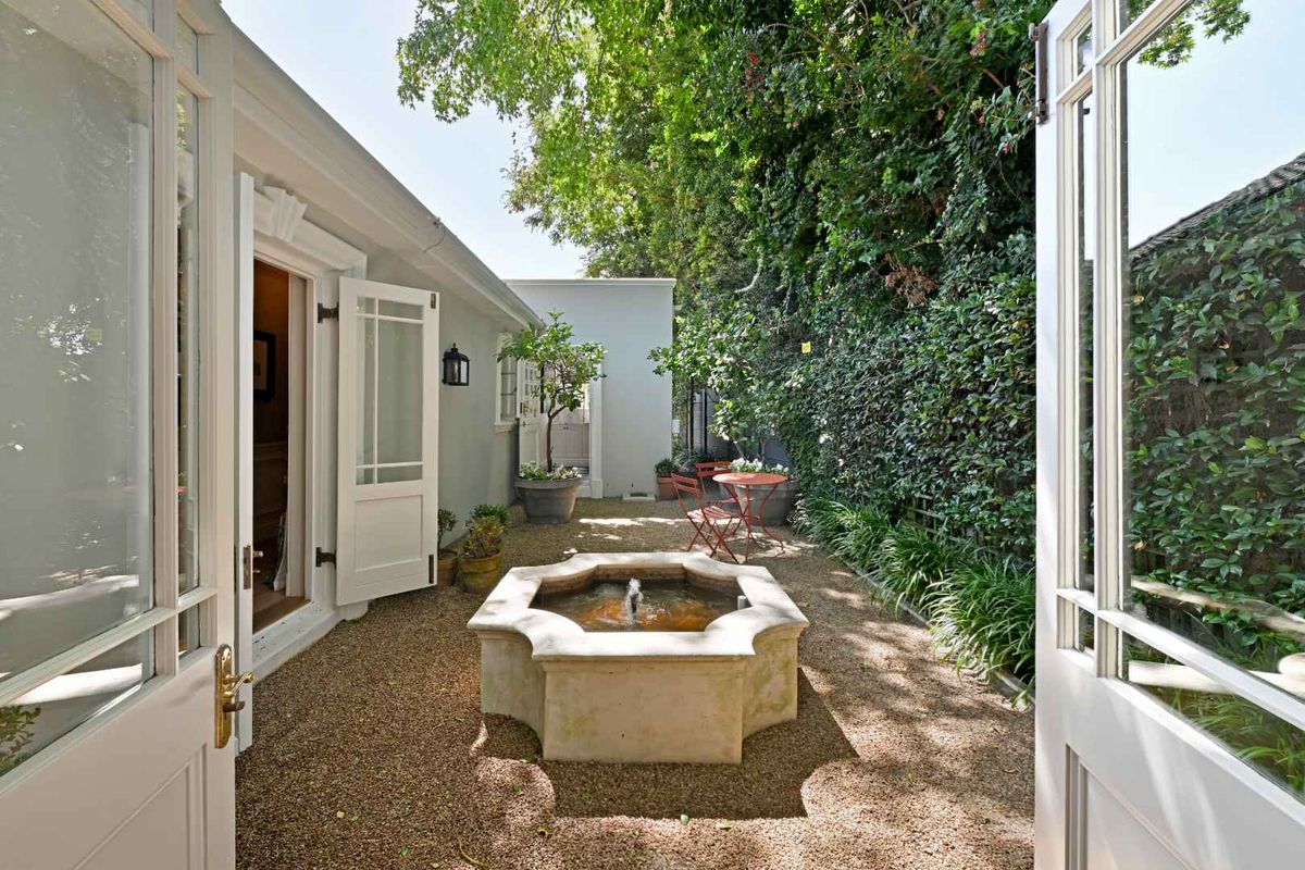 A charming, private courtyard garden featuring a central stone fountain, gravel flooring, and lush perimeter greenery, accessible through double glass doors.