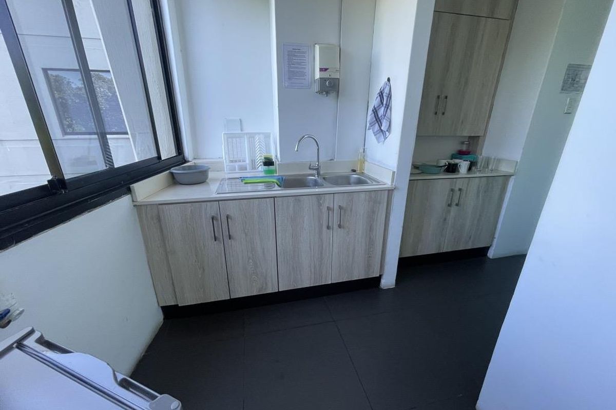 A functional office kitchenette featuring light wood-grain cabinetry, a stainless steel double sink, and dark floor tiling.