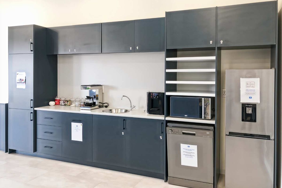 A modern office kitchenette featuring dark grey cabinetry, integrated shelving, a stainless steel sink, and space for built-in appliances.