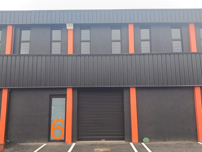 Modern industrial unit facade featuring dark metal cladding, orange structural accents, roller shutter doors, and dedicated parking bays.