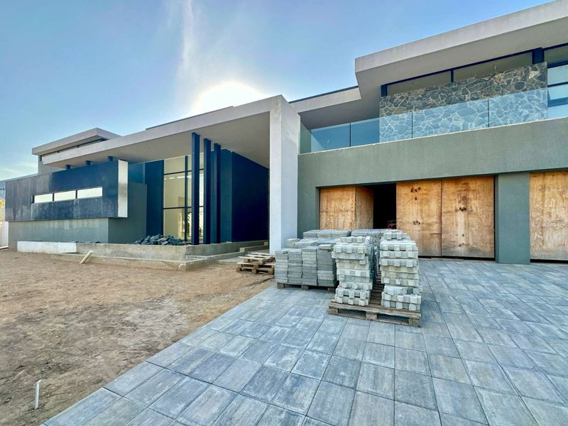 Modern architectural home exterior under construction, featuring stone cladding, glass balcony railings, and paved driveway area.