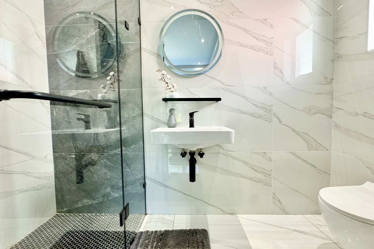Modern bathroom featuring marble-look wall tiles, a glass-enclosed shower, wall-mounted basin with matte black fixtures, and a contemporary circular LED mirror.
