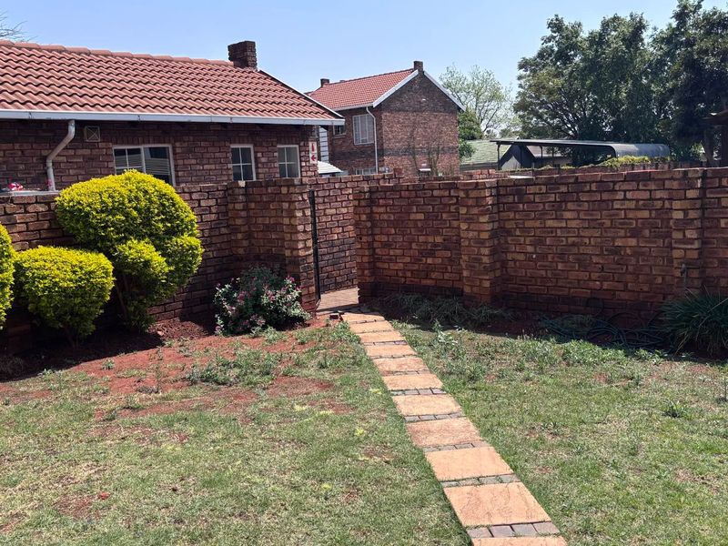 A private rear garden area featuring a face brick boundary wall, tiled roof structure, and a paved walkway leading to a metal gate.