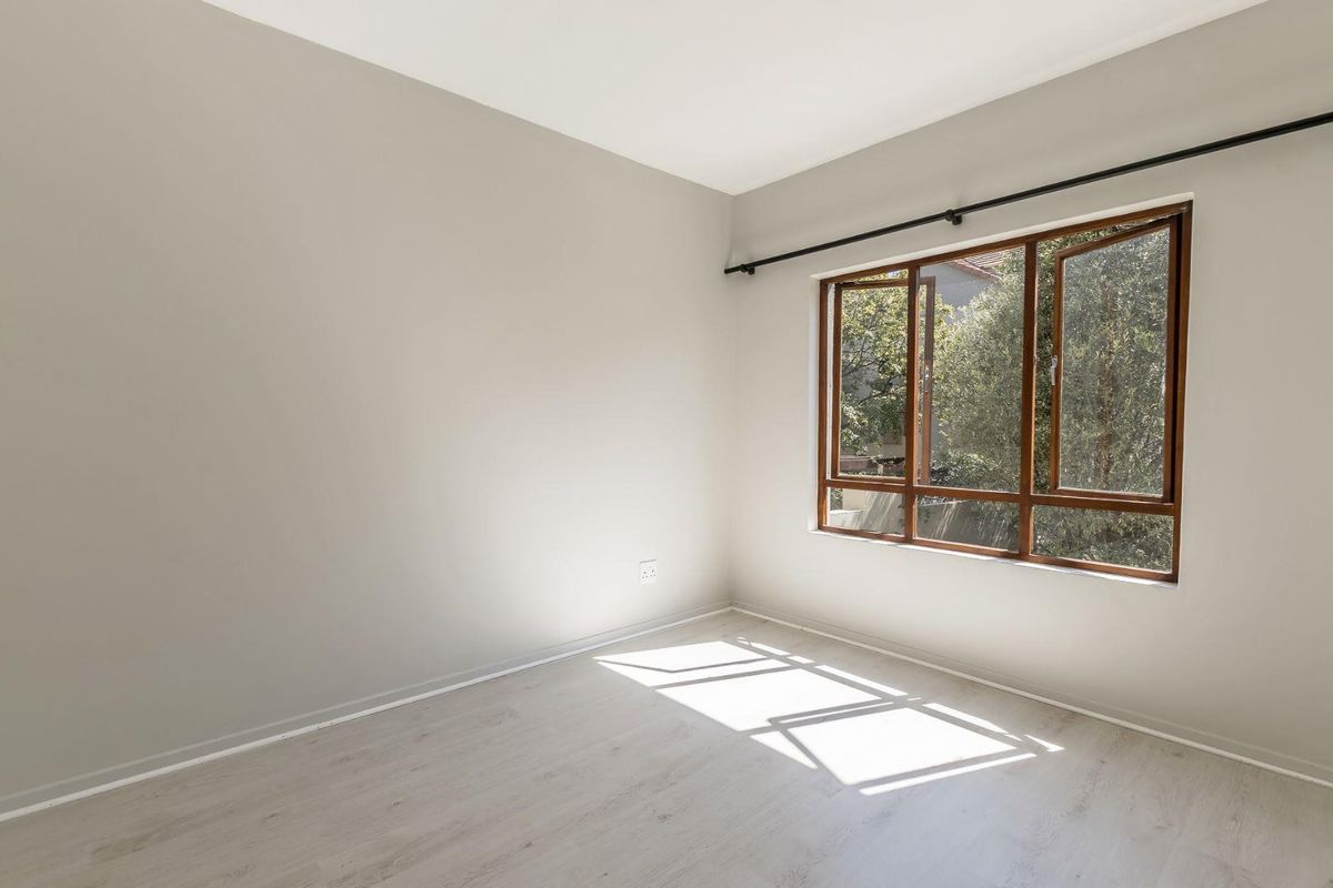 A bright, empty bedroom featuring light-toned laminate flooring, neutral walls, and a large window with wooden frames.