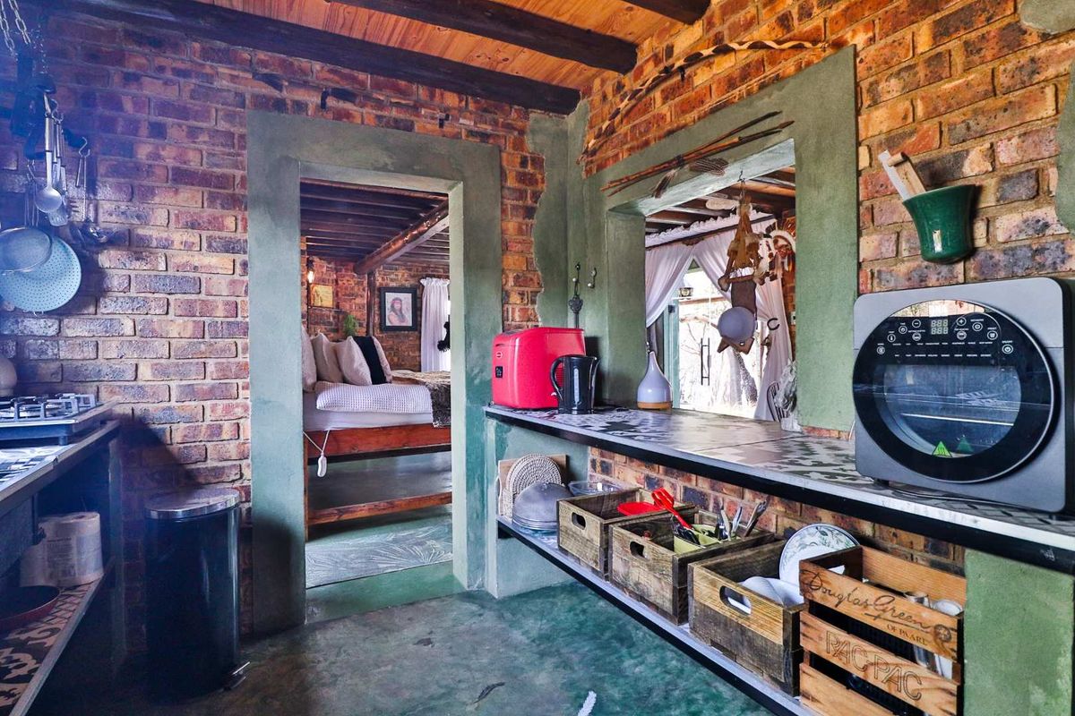 Rustic kitchen area featuring exposed brick walls, timber ceiling beams, and a tiled countertop, with an open doorway leading into the adjacent bedroom.