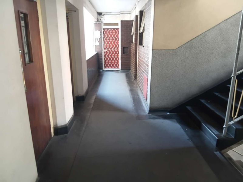 A communal apartment hallway featuring a staircase, brick wall accents, and a security gate, showing signs of wear and dated finishes.