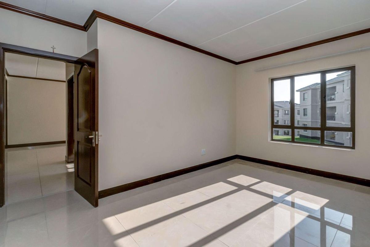A bright, empty bedroom featuring tiled flooring, dark wood trim, and a window overlooking the apartment complex grounds.