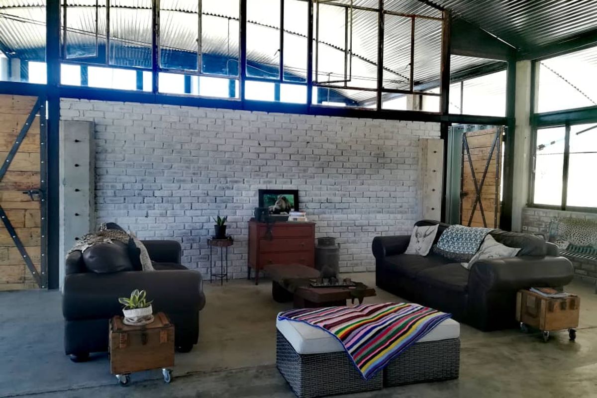 Spacious industrial-style living area featuring exposed brick walls, high vaulted corrugated metal ceiling, and large steel-framed windows providing ample natural light.