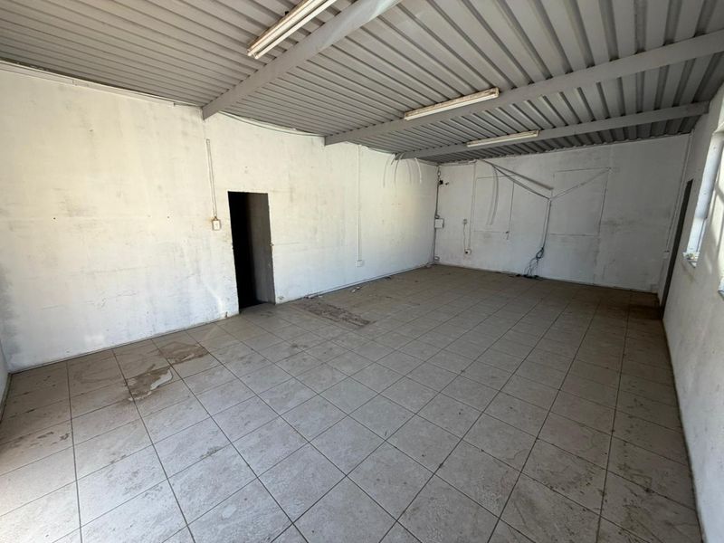 An empty, utilitarian commercial space featuring tiled flooring, exposed corrugated metal ceiling, and basic fluorescent lighting, suitable for storage or workshop use.