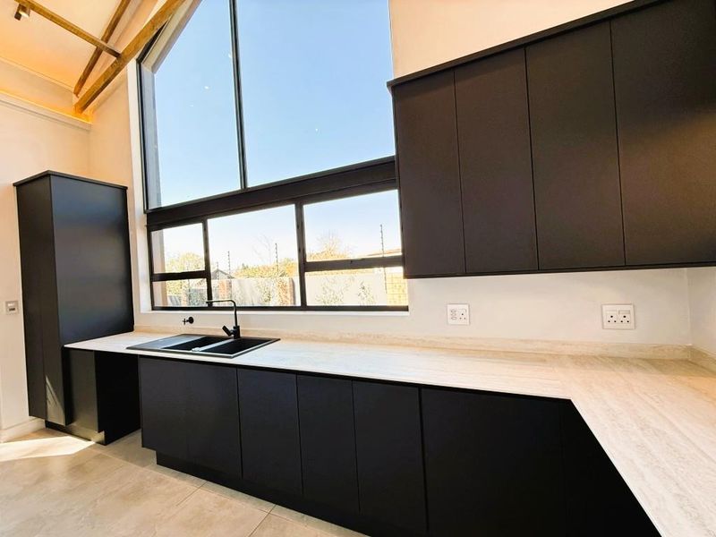 Modern kitchen featuring sleek matte black cabinetry, stone-look countertops, a black sink, and a striking vaulted ceiling with exposed timber beams and large windows.